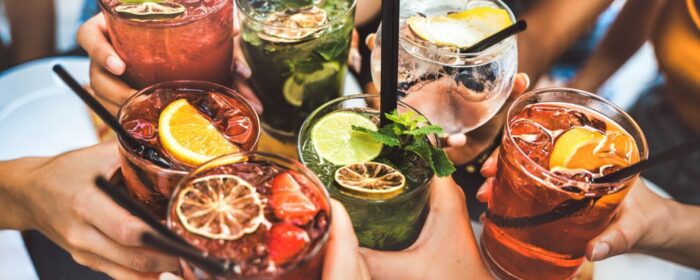 A vibrant, high-angle shot of seven people holding up a variety of colorful cocktails with garnishes like mint, citrus slices, and berries. The image conveys a festive and social atmosphere, serving as an invitation to the happy hour event at IBC 2025.